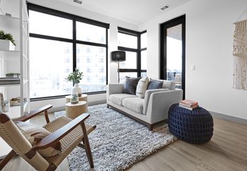 A living room with a grey couch, a wooden chair, a white rug, and a large window.