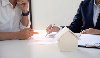 A person in a white shirt is signing a document with a pen while another person in a blue suit is pointing at the document.