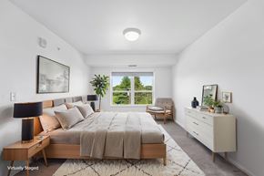Faribault apartment for rent - A bedroom with a large bed, a chair, and a window overlooking a garden.