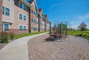 Hugo apartment for rent - A playground with a swing set is located in front of a building with a paved walkway.