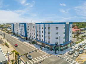 Bradenton apartment for rent - A large white and blue building is situated on a street corner with cars and a traffic light.
