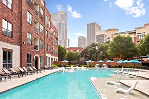 Houston apartment for rent - A pool surrounded by chairs and umbrellas in front of a brick building.