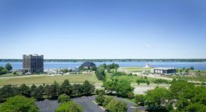Muskegon apartment for rent - A large building sits on the left side of a body of water, with a parking lot and trees in the foreground.