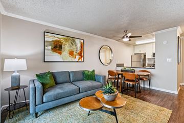 Alameda apartment for rent - A living room with a grey couch, a painting on the wall, a round mirror, and a dining area in the background.