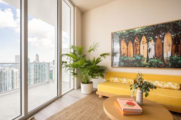Miami apartment for rent - A living room with a yellow couch, a painting on the wall, and a view of the city through the windows.