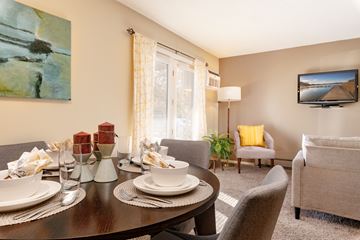 Fridley apartment for rent - A dining table set with plates and glasses is in the foreground of a living room with a couch and television.