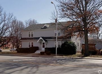 Bartlesville apartment for rent - A house with a grey roof and a white door is surrounded by trees and bushes.