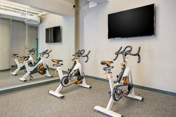 Three stationary bikes are lined up in a row in a room with a wall-mounted tv.