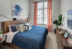 Denver apartment for rent - A bedroom with a blue bedspread and pillows, a wooden headboard, and a window with orange curtains at Regatta Sloans Lake Apartments, Denver, 80204