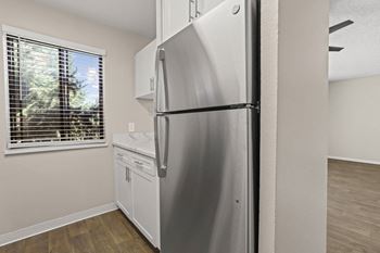 A stainless steel refrigerator in a kitchen with wooden flooring and a window with blinds.
