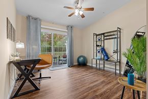 Zeeland apartment for rent - A room with a wooden floor, a ceiling fan, a table with a blue bottle, a chair, a window with a view of trees, a gym equipment, and a potted plant.
