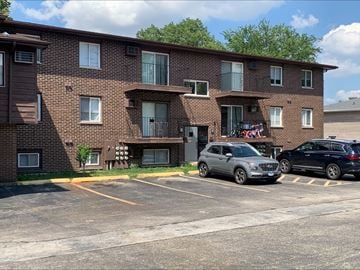 Dekalb apartment for rent - A parking lot in front of a brown brick apartment building with two cars parked.