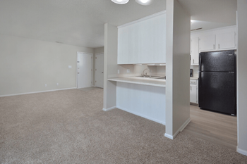 An empty living room with a kitchen and a black refrigerator