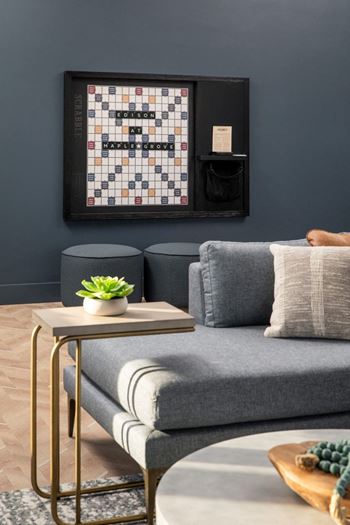 A living room with a grey couch, a small table with a plant and a book, and a framed board game on the wall.