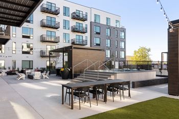A patio with a wooden table and chairs and a pool in front of an apartment building