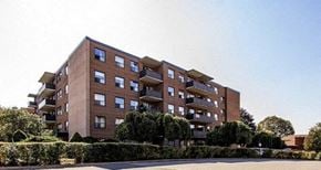 North York apartment for rent - A large brick building with balconies and windows is surrounded by green bushes.