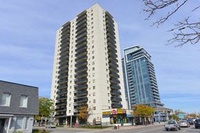 Mississauga apartment for rent - A tall beige building with balconies is in the middle of a street with cars and other buildings.