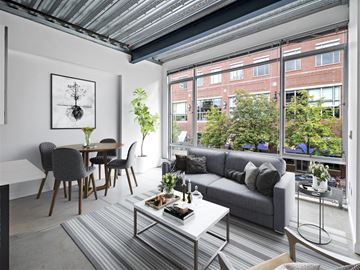 Cambridge apartment for rent - A living room with a grey couch, a grey and white coffee table, a grey dining table with grey chairs, a grey rug, a white wall with a framed picture, a window with a view of a street and buildings,