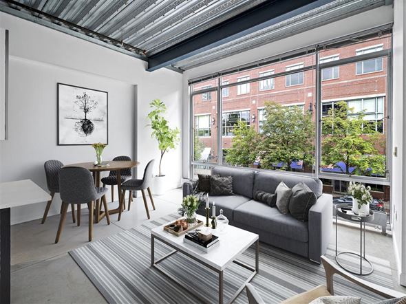 Cambridge apartment for rent - A living room with a grey couch, a grey and white coffee table, a grey dining table with grey chairs, a grey rug, a white wall with a framed picture, a window with a view of a street and buildings,