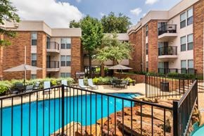 Dallas apartment for rent - A swimming pool with a waterfall surrounded by lounge chairs and a couple of tables, chairs and umbrellas at Cambridge Court Apartments Entryway in Lake Highlands, Dallas, TX.