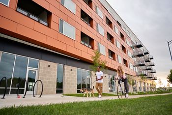 A woman riding a bicycle next to a man walking a dog in front of a modern building.