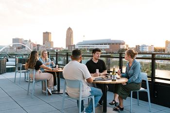 Four people are sitting at a table on a rooftop with a city skyline in the background.