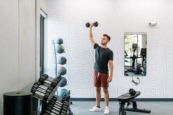 A man lifting dumbbells in a gym with a mirror and weight rack.