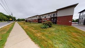 Fredericton apartment for rent - A long building with a red brick exterior sits next to a grassy area.