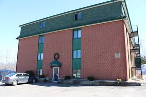 Fredericton apartment for rent - A red brick building with a green roof and a white car parked in front.