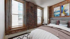 Denver apartment for rent - A bedroom with a large bed and an exposed brick wall with 2 large floor-to-ceiling windows.