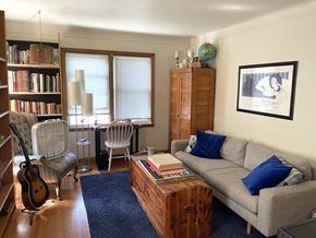 St. Paul apartment for rent - A living room with a grey couch, a wooden bookshelf, a chair, a guitar, a cabinet, a table, a window with blinds, a rug, a blue pillow, a picture frame, and a globe.