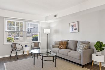 A living room with a grey couch, a glass table, and a window with a view of the street