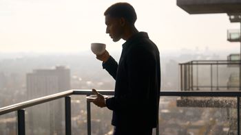 A man in a black jacket is standing on a balcony holding a white cup.