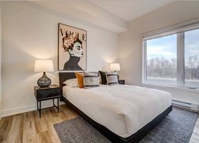 Gatineau apartment for rent - A bedroom with a large bed, a side table with a vase, and a large painting of a woman's face on the wall at Champlain Village Urbain Apartments , Gatineau, J9H 0K9
