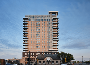 Gatineau apartment for rent - A tall building with a mix of brick and glass facade stands in front of a smaller house and a tree. at Le ColumbiaApartments , Gatineau, J8Y 0A8