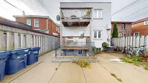 Toronto apartment for rent - A grey building with a balcony is in the middle of a courtyard with blue bins and a fence.