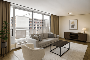 Hamilton apartment for rent - A living room with a grey couch, a white chair, a brown coffee table, and a large window with a view of a building outside.