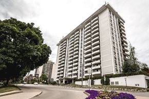 Hamilton apartment for rent - A tall residential building with balconies is surrounded by trees and a street with a flower bed.