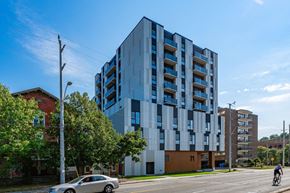 Dundas apartment for rent - A modern multi-story building with blue and white balconies is situated on a street corner with a car and a cyclist in the foreground.