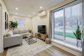 Hamilton apartment for rent - A living room with a grey couch, a coffee table, a flat screen TV, and a large window with a view of a green lawn.