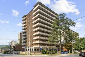 Hamilton apartment for rent - A large brown building with many windows and balconies is surrounded by trees and a street with cars.
