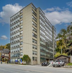 Hamilton apartment for rent - A tall beige building with many windows is surrounded by trees and parked cars.