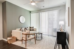 Decatur apartment for rent - A living room with a white couch, a wooden chair, a round mirror, a small table, a lamp, and a window with blinds.