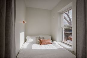 Detroit apartment for rent - A bedroom with a white bed and a window with a view of a cityscape.