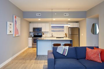Kansas City apartment for rent - A blue couch with a red pillow is in the foreground of a modern kitchen and living room.