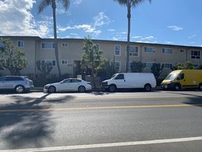 Los Angeles apartment for rent - A white car is parked on the side of the road in front of a building with a yellow van and a white van parked behind it.