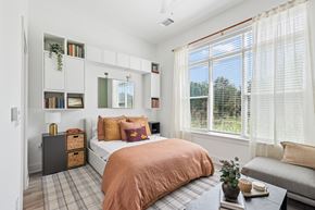 Charlotte apartment for rent - A bedroom with a bed, a window with blinds, a nightstand, a bookshelf, and a plant on a table at Link Apartments® CYKEL Apartments, Charlotte, NC, 28208