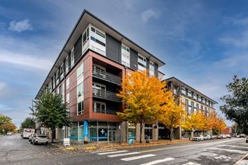 Portland apartment for rent - A modern building with a red and grey facade stands on a street corner with trees showing autumn colors.