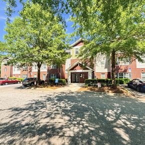 Greenville apartment for rent - A building with a red brick facade and a large front yard with trees and a gravel area.