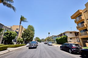 Los Angeles apartment for rent - Cars driving down a street with palm trees and buildings on either side.